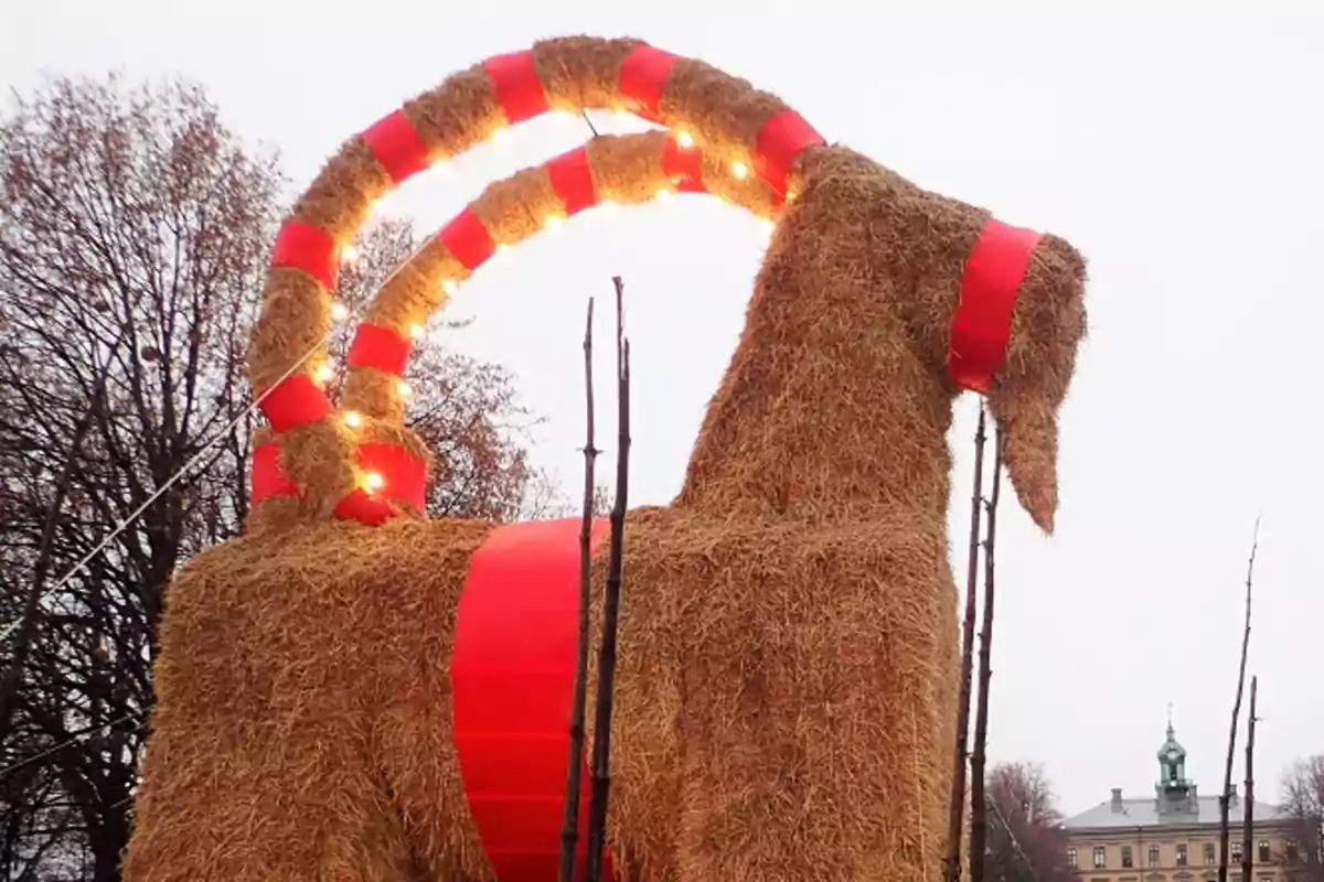 Una gran figura de cabra hecha de paja con cintas rojas y luces en un entorno al aire libre. tradición navideña en Suecia.