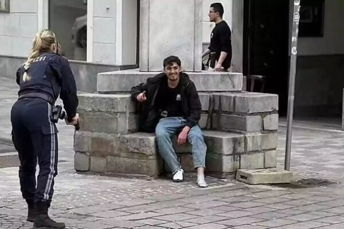 A police officer points a gun at a man sitting on a stone structure on an urban street while another person walks nearby.