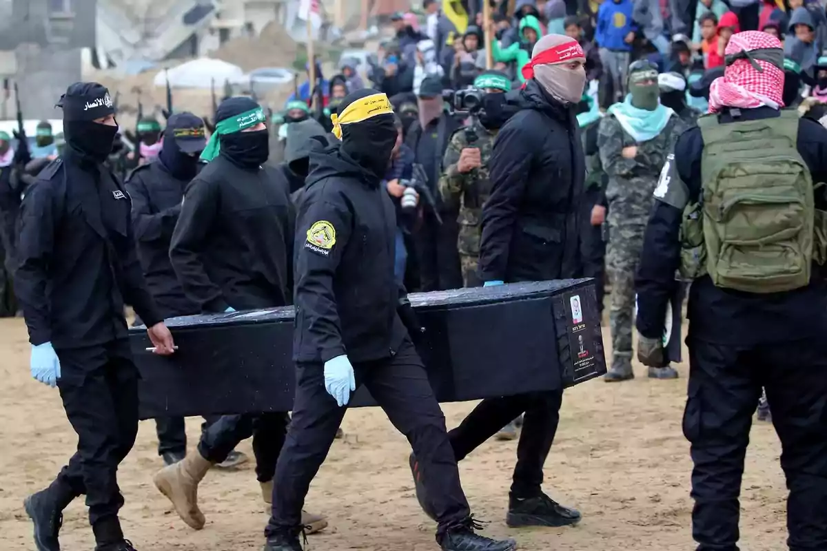 Un grupo de personas vestidas de negro y con el rostro cubierto lleva un ataúd en un entorno al aire libre, rodeados de más personas y algunos con cámaras.