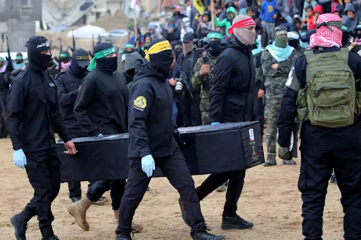 Un grupo de personas vestidas de negro y con el rostro cubierto lleva un ataúd en un entorno al aire libre, rodeados de más personas en el fondo.