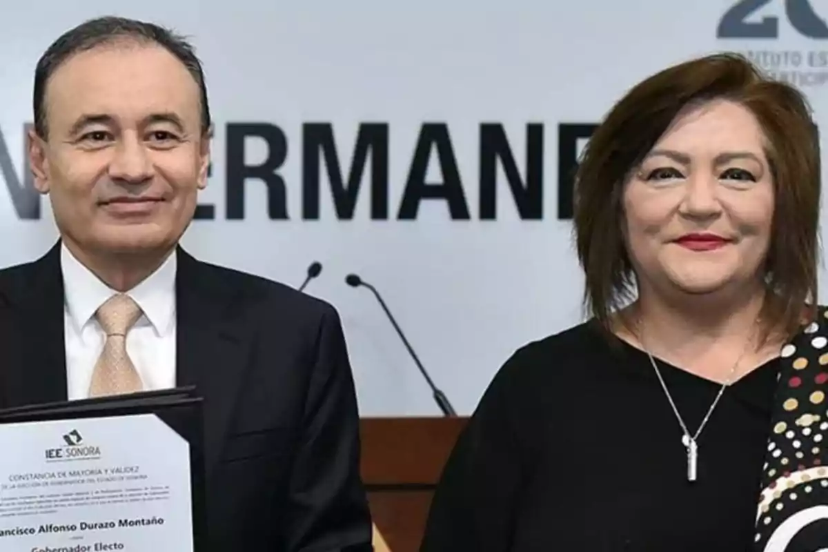 Two people pose together at a formal event, one of them holding a framed document.