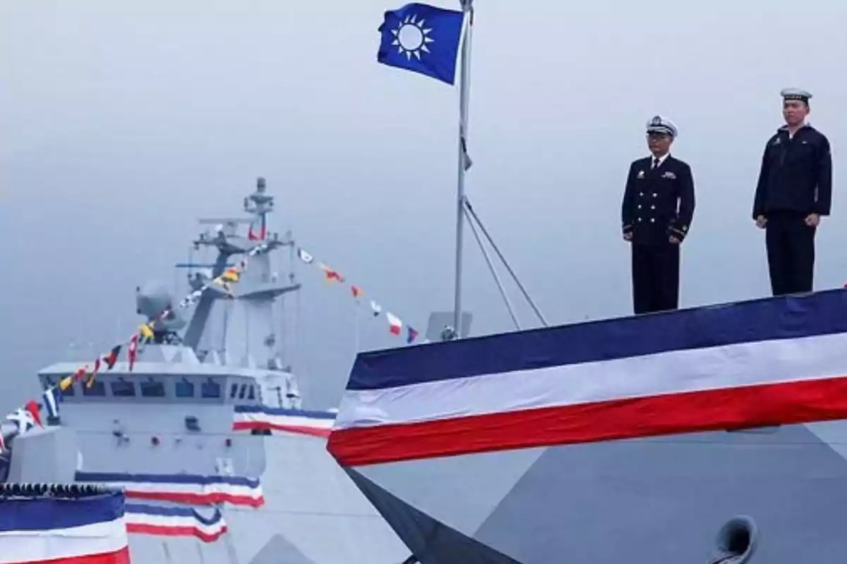 Dos oficiales navales de pie en la cubierta de un barco decorado con banderas, con otra embarcación al fondo y una bandera azul ondeando en el mástil.