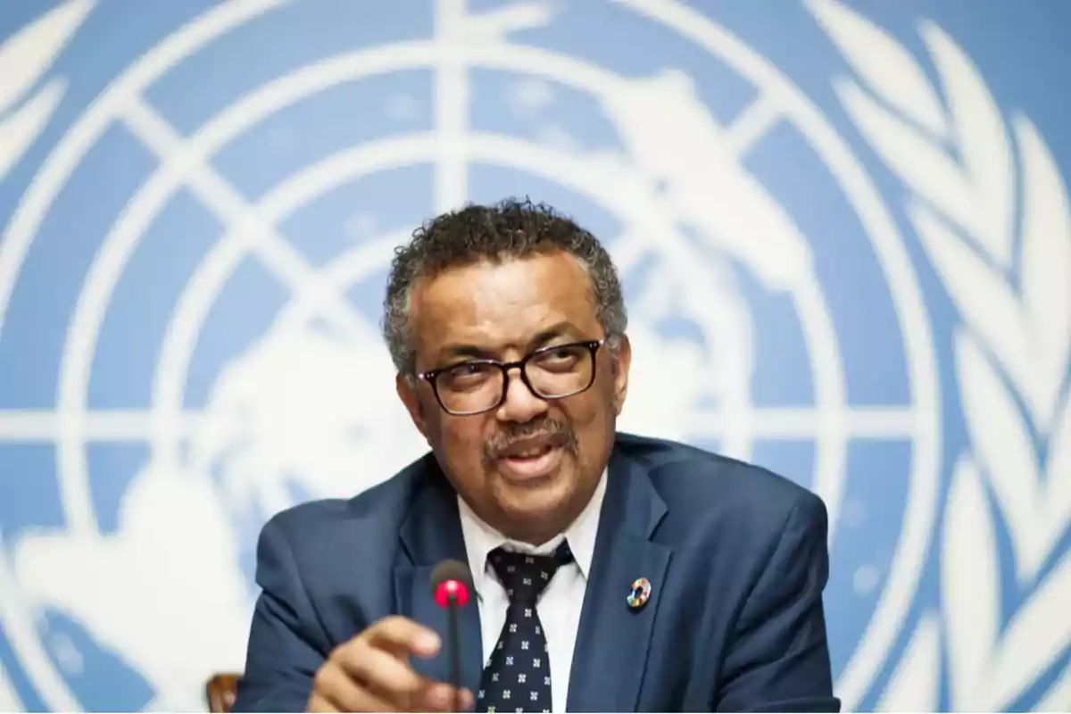 Un hombre con gafas y traje oscuro habla frente a un micrófono con un fondo azul que tiene un logotipo circular.