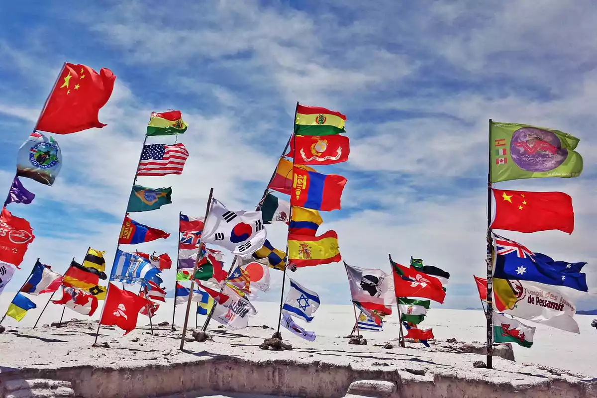 A group of flags from different countries waves in the wind in a desert landscape under a blue sky.