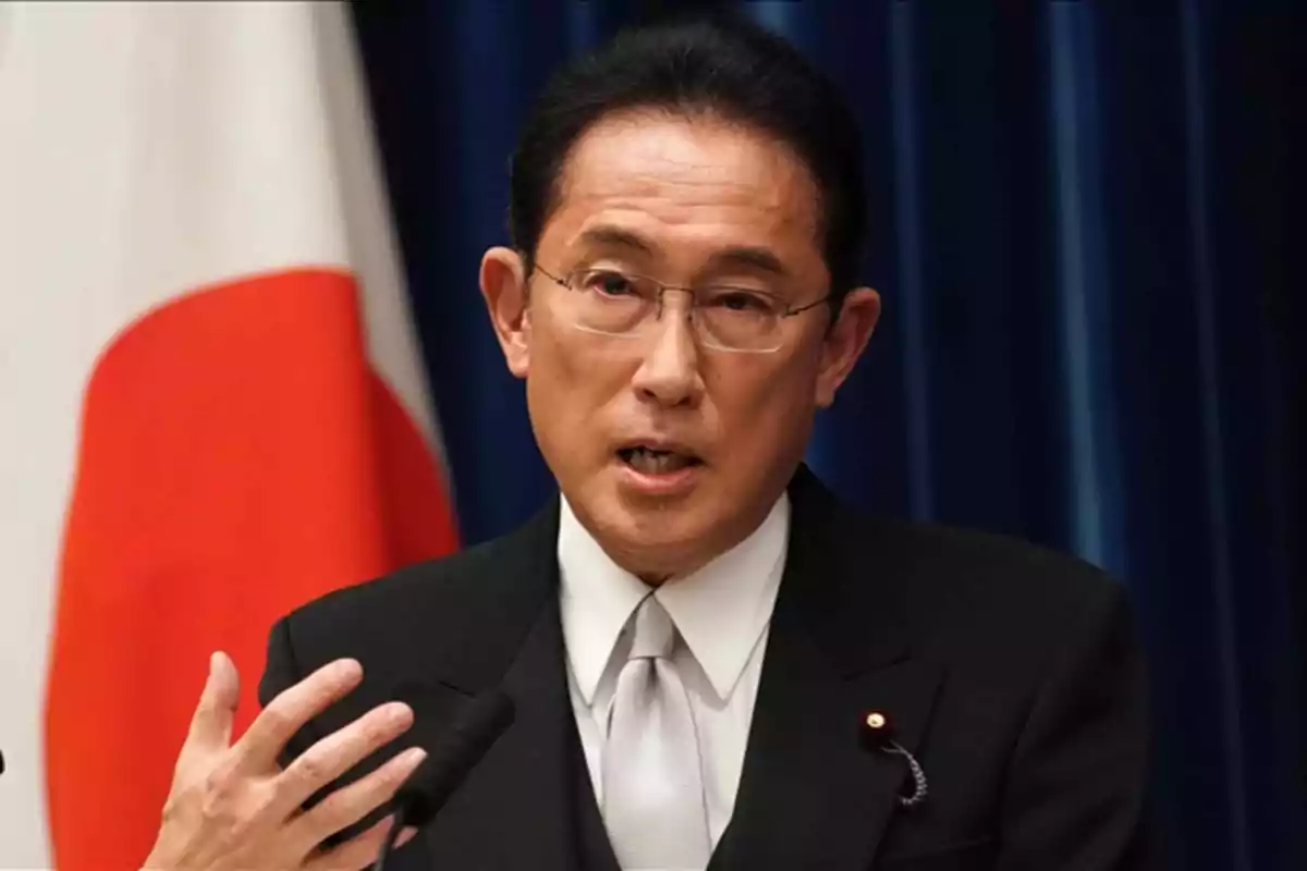 Un hombre con gafas y traje oscuro hablando frente a un micrófono con una bandera de Japón en el fondo.