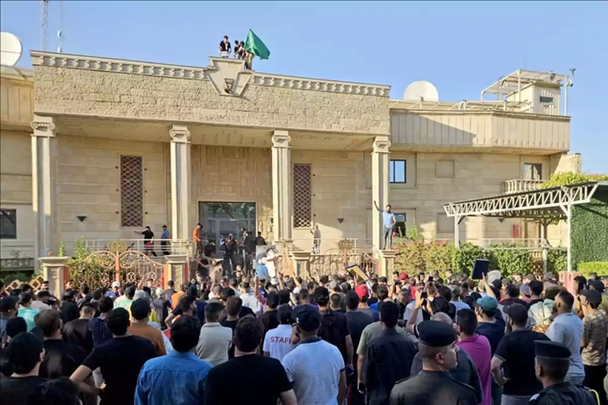 Una multitud se reúne frente a un edificio con columnas, mientras algunas personas están en el techo ondeando una bandera verde.