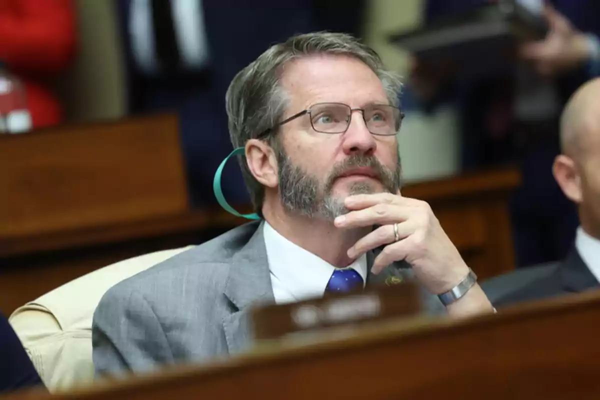 Un hombre con barba y gafas, vestido con traje gris y corbata azul, está sentado en una sala, apoyando su barbilla en su mano mientras parece estar escuchando atentamente.