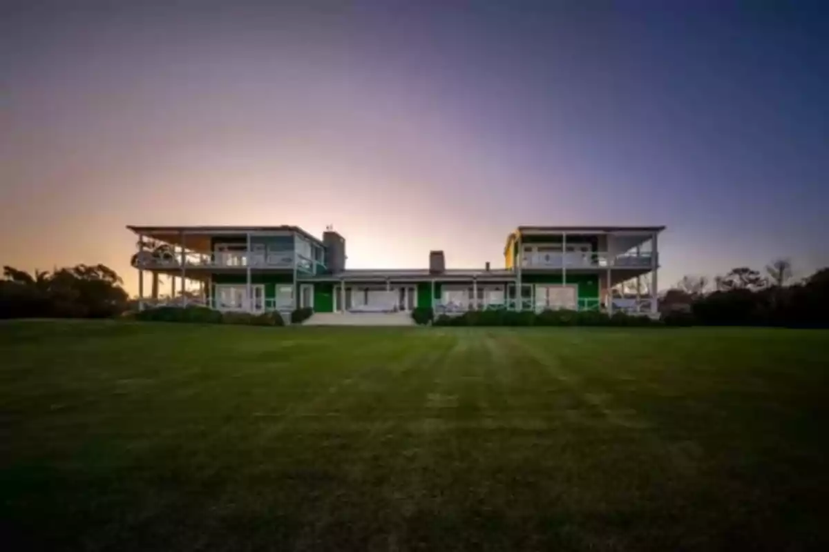 Una casa moderna de dos pisos con amplios ventanales y balcones rodeada de un extenso césped al atardecer.