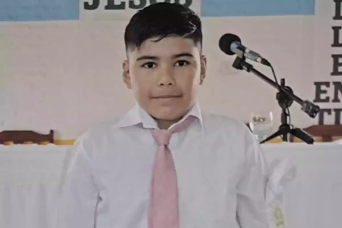 Niño con camisa blanca y corbata rosa de pie frente a un micrófono.