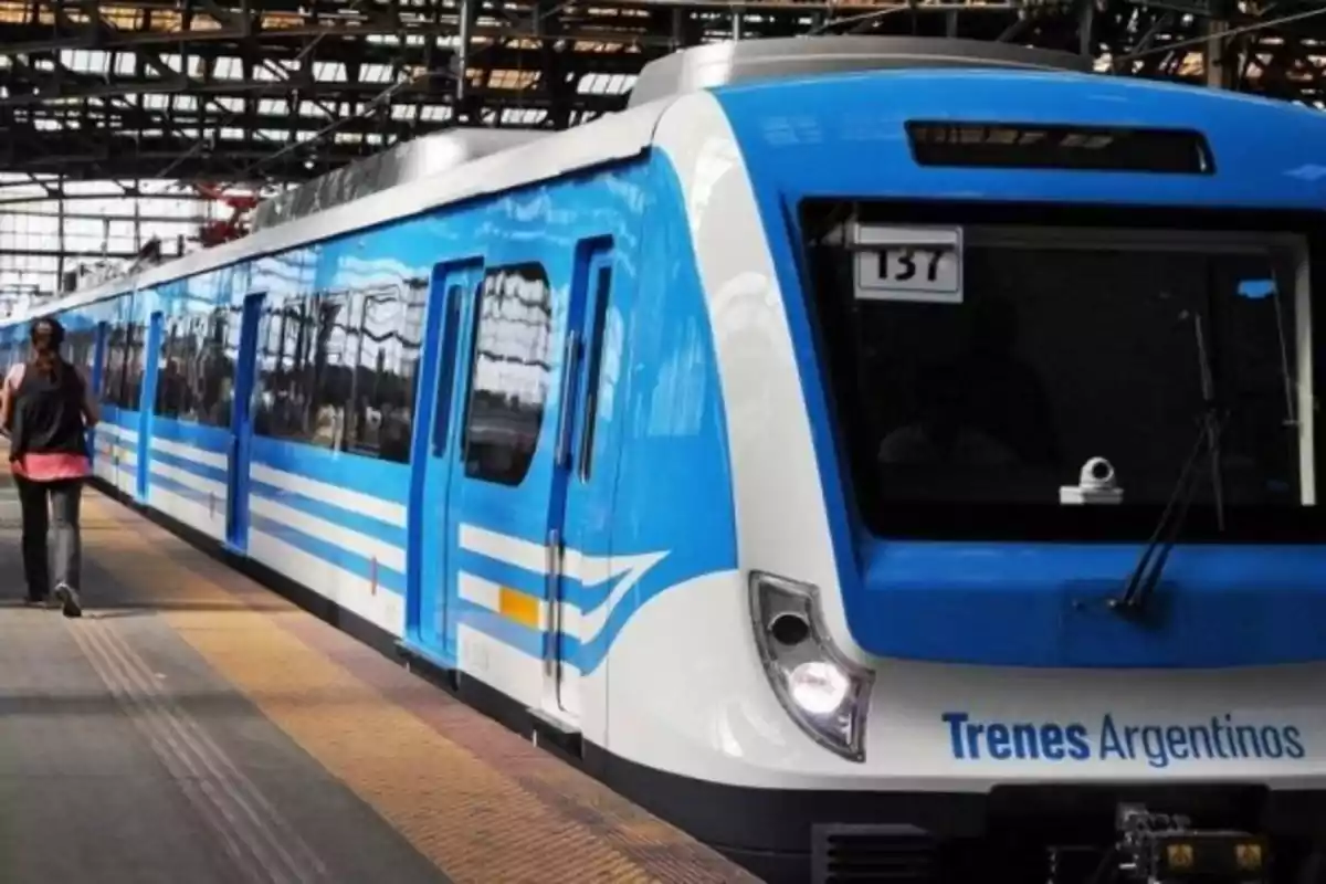 Un tren azul y blanco de Trenes Argentinos en una estación con una persona caminando por el andén.