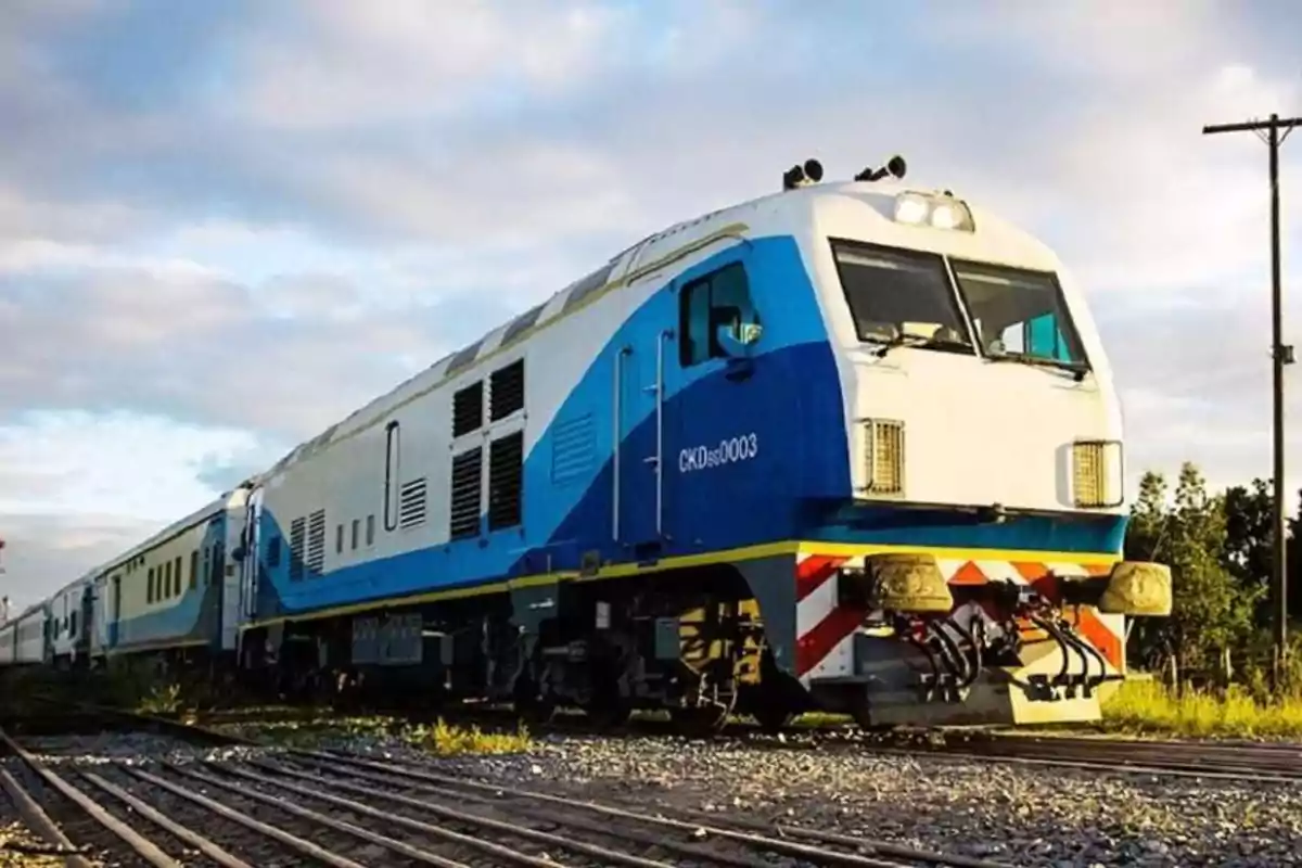 Un tren azul y blanco sobre las vías en un paisaje al atardecer.