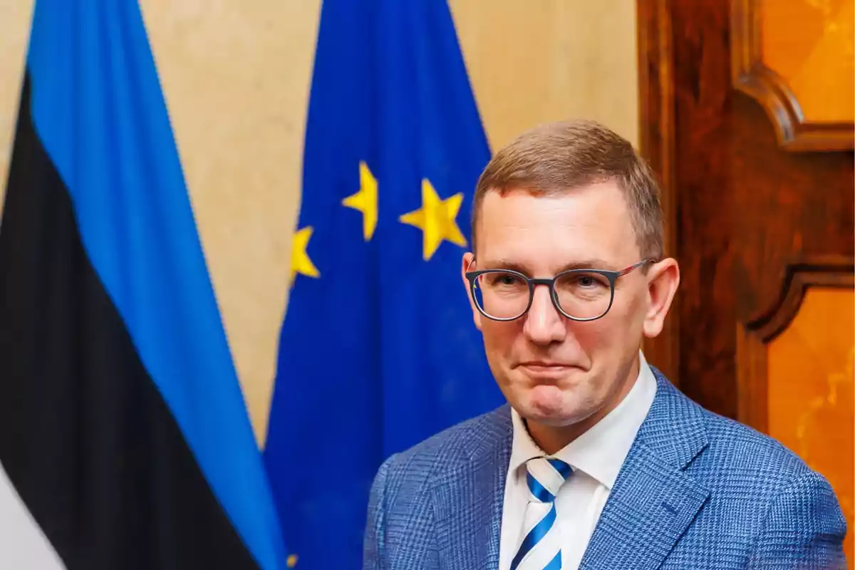 Un hombre con gafas y traje azul está de pie frente a las banderas de Estonia y la Unión Europea.