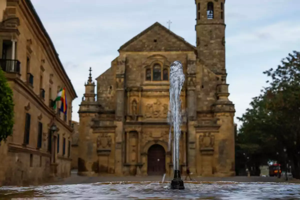 Una fuente de agua en primer plano con una iglesia histórica de fondo en una plaza de un pueblo de españa donde ofrecen trabajo y estadia gratis.