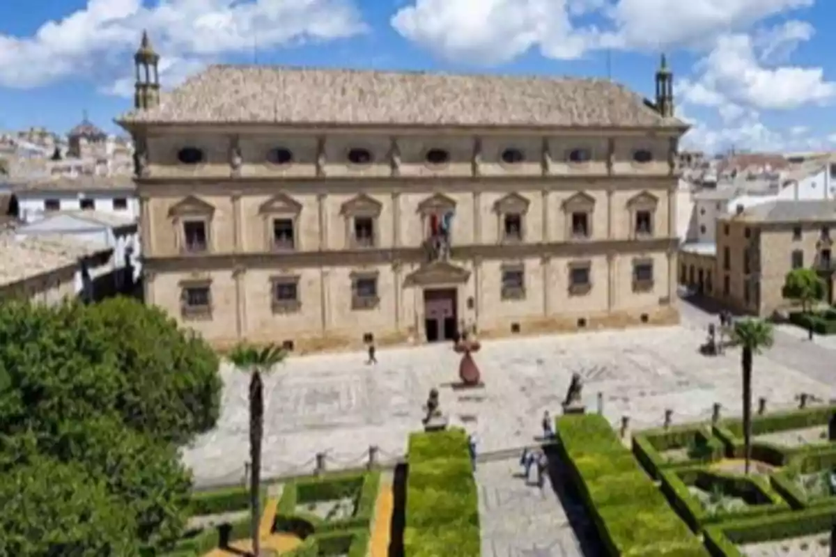 Edificio histórico con fachada de piedra y jardín con setos y palmeras en primer plano bajo un cielo azul con nubes en un pueblo de España donde ofrecen trabajo y vivienda gratis.