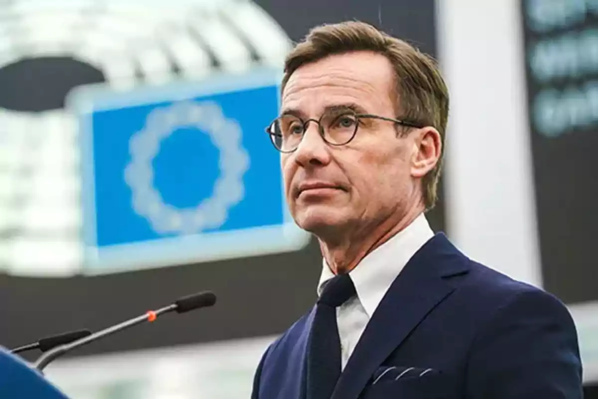 Un hombre con gafas y traje oscuro hablando frente a un micrófono con un fondo que incluye un símbolo azul.