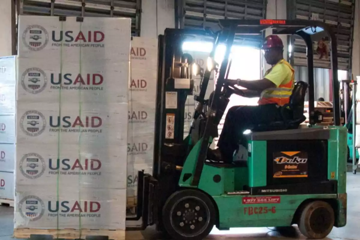 Un trabajador operando una carretilla elevadora transporta cajas con el logo de USAID en un almacén.