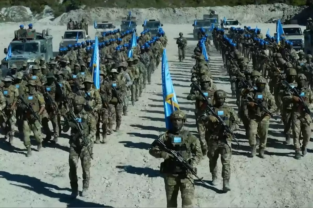 Soldados marchando en formación con cascos azules y banderas azules en un terreno arenoso.