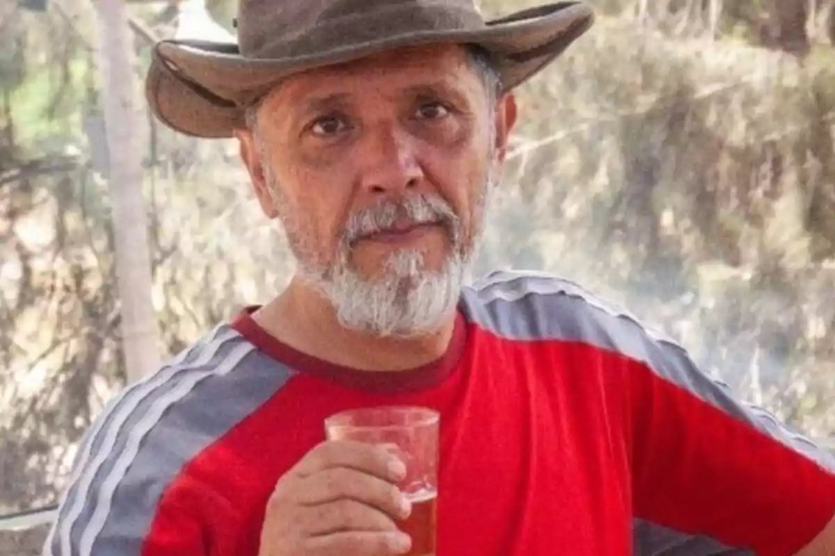 Un hombre con barba y sombrero sostiene un vaso mientras viste una camiseta roja con rayas grises en las mangas, con un fondo de árboles desenfocado.