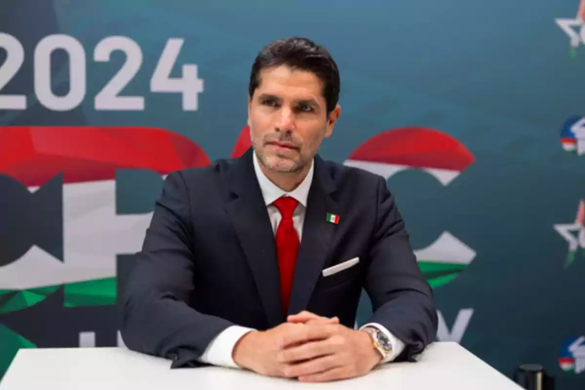 Un hombre con traje oscuro y corbata roja está sentado frente a un fondo con el número 2024 y colores verde, blanco y rojo.