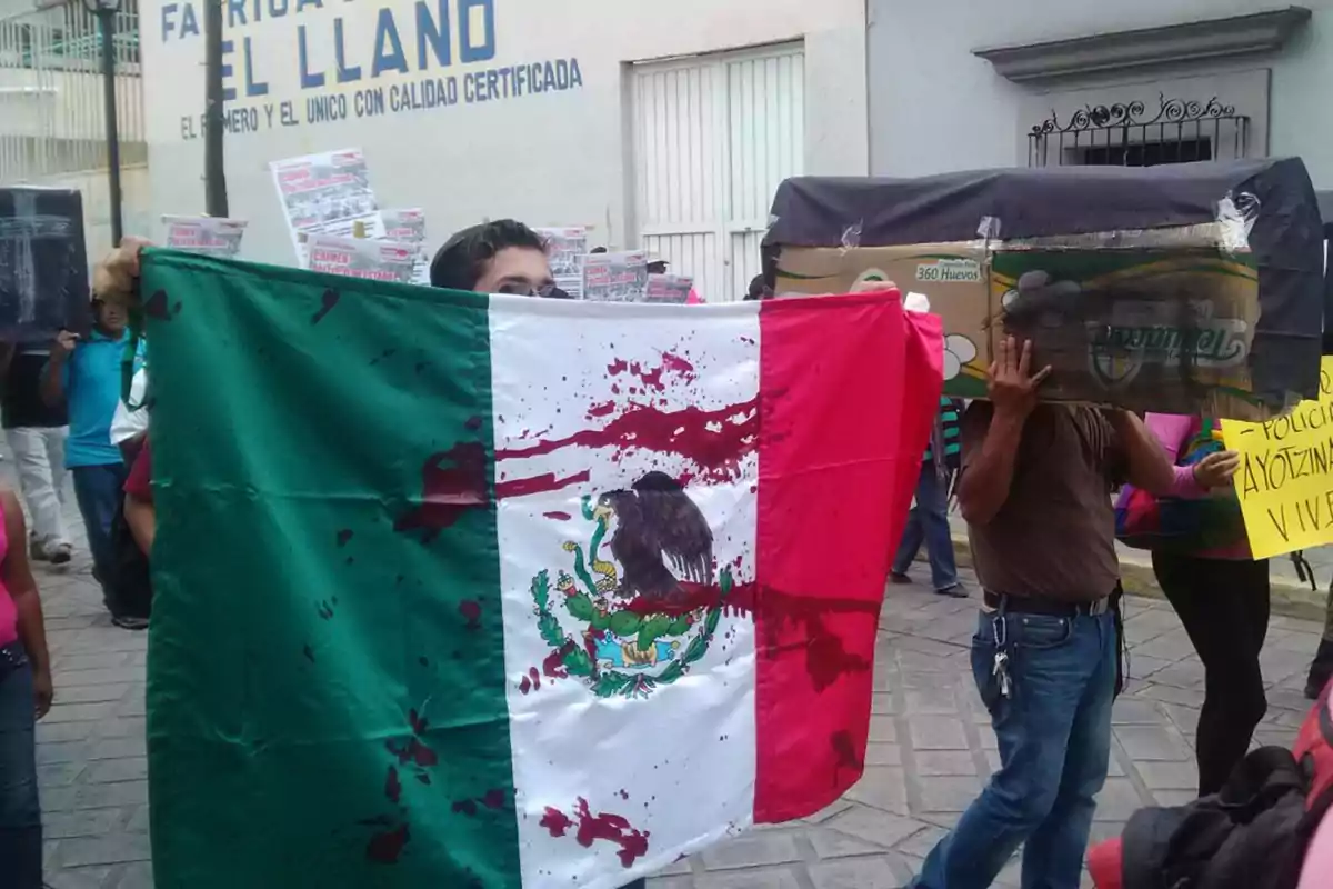 A person holds a Mexican flag with red stains at a street protest, surrounded by other people carrying signs and boxes.