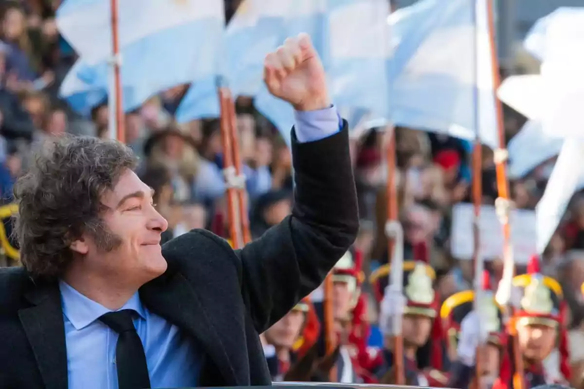 Un hombre sonriente levanta el puño en un evento con banderas argentinas y guardias de fondo.