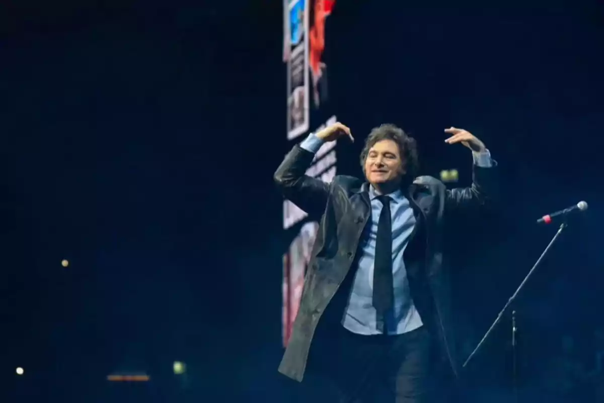 Un hombre con abrigo y corbata gesticula con las manos en un escenario iluminado.