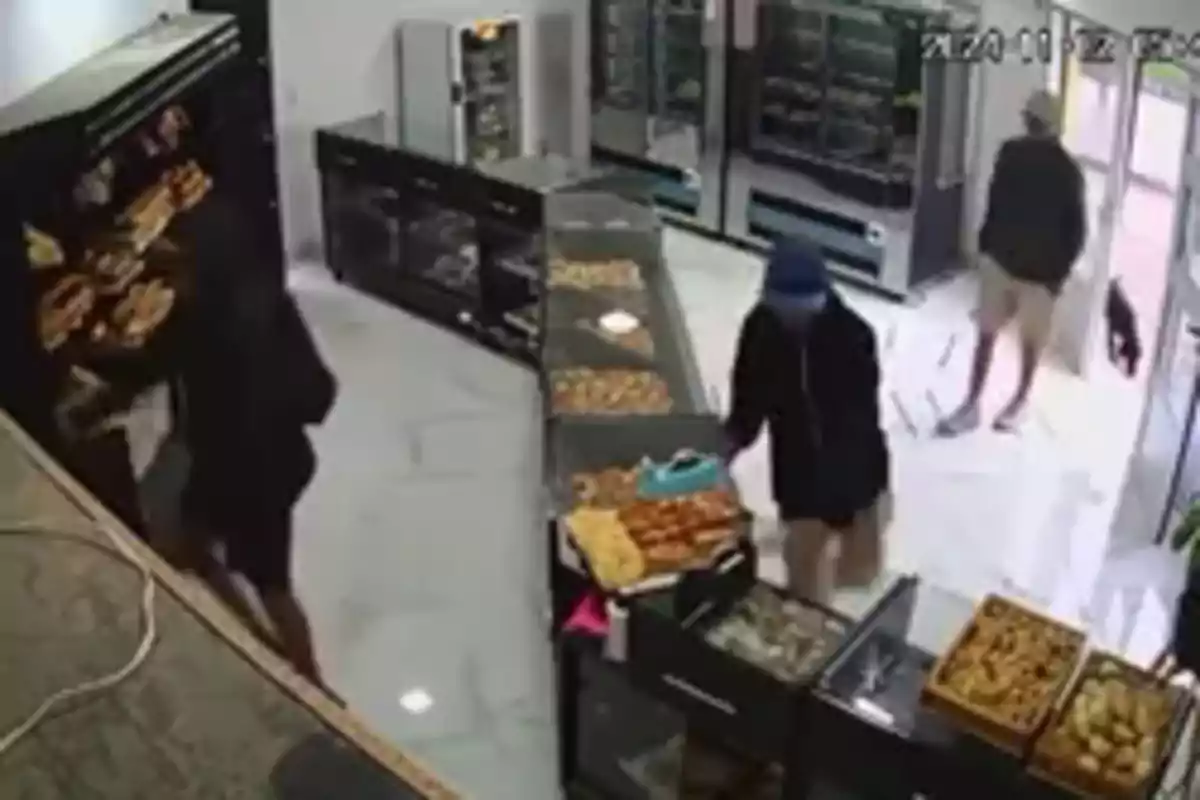 Tres personas con ropa oscura y capuchas están dentro de una panadería o tienda de alimentos con vitrinas llenas de productos horneados.