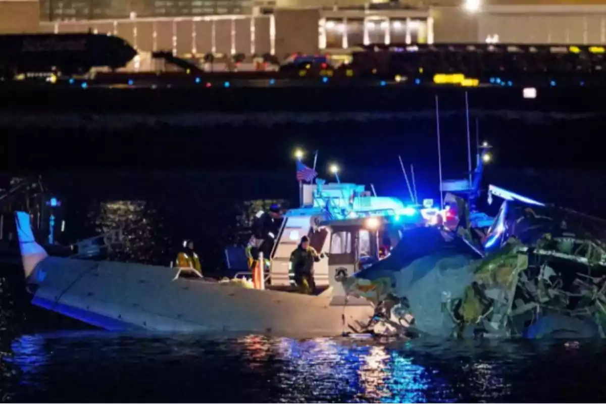 Una embarcación de rescate iluminada con luces azules se encuentra junto a los restos de un avión en el agua durante la noche, con un edificio iluminado al fondo.