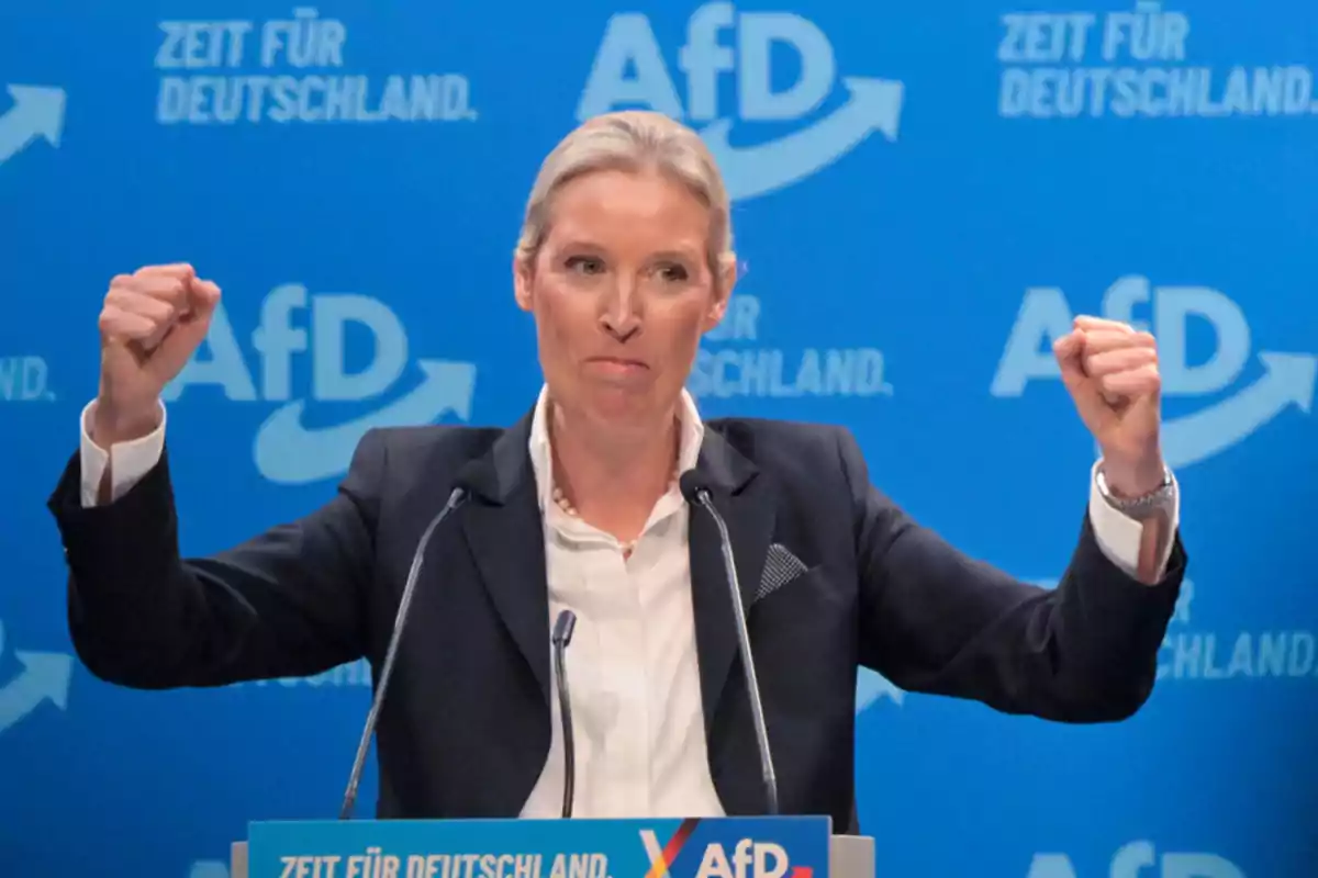 Una persona en un podio con fondo azul y logotipos de AfD, levantando los brazos en un gesto de triunfo.