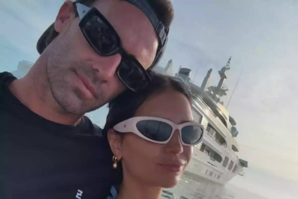 Una pareja con gafas de sol posa frente a un yate en el agua.