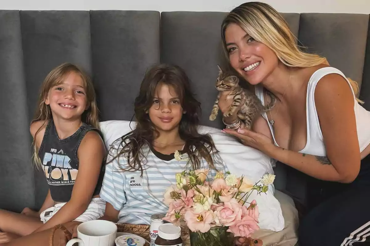 Tres personas sonríen mientras una de ellas sostiene un gatito, con una bandeja de desayuno y un ramo de flores frente a ellas.