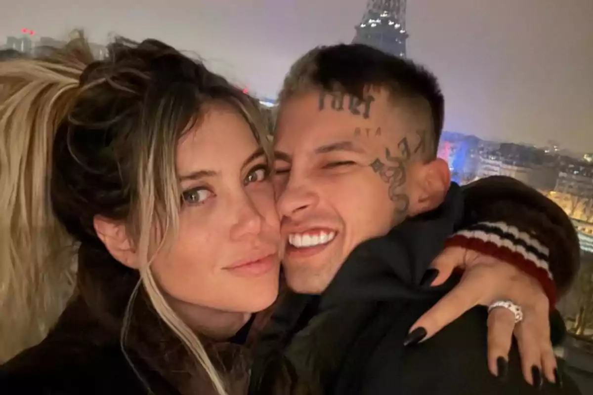 Una pareja sonriente se abraza frente a un paisaje urbano nocturno con una torre iluminada al fondo.