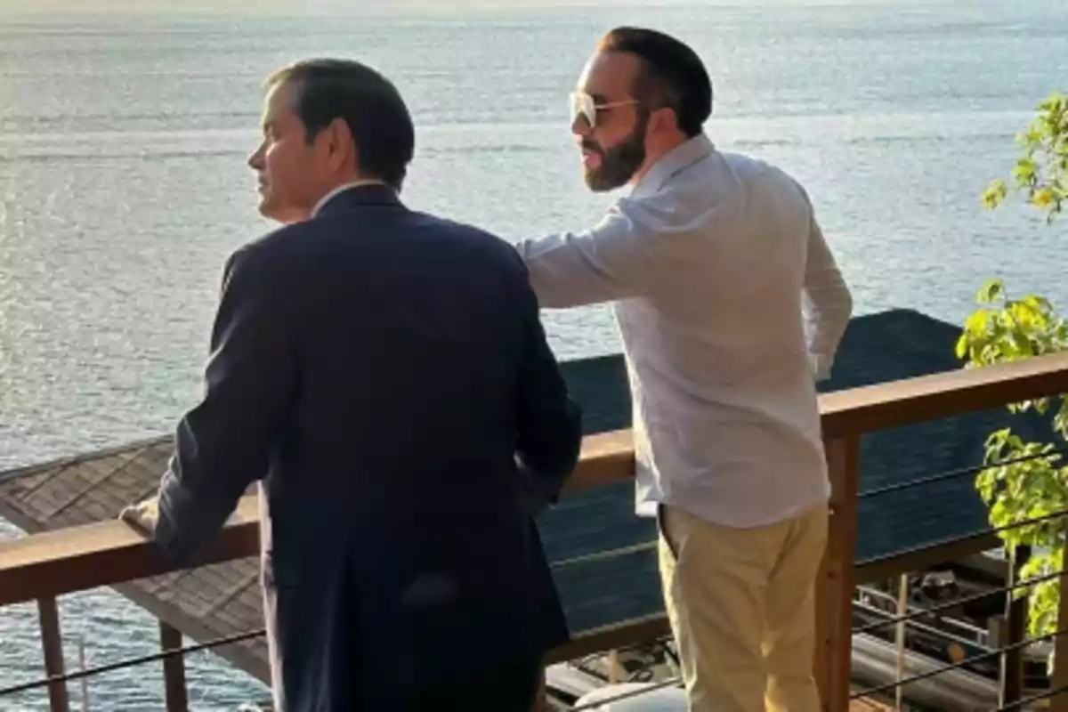 Dos personas de pie en un balcón con vista al mar en un día soleado.