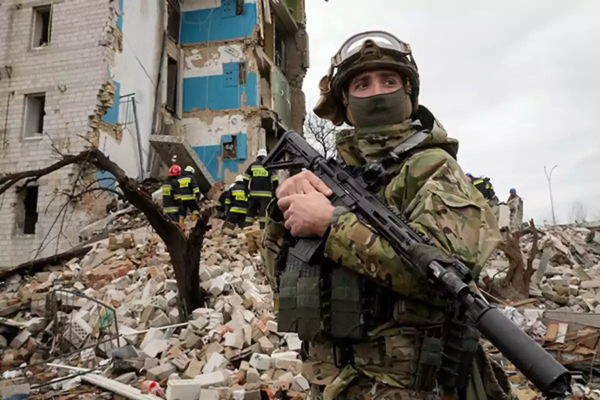 Un soldado armado con uniforme de camuflaje está de pie frente a un edificio dañado mientras los rescatistas trabajan entre los escombros.