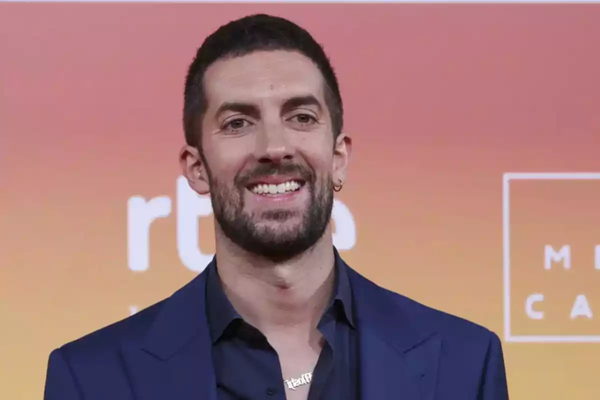 Hombre con barba y cabello corto sonriendo en un evento con fondo degradado naranja.