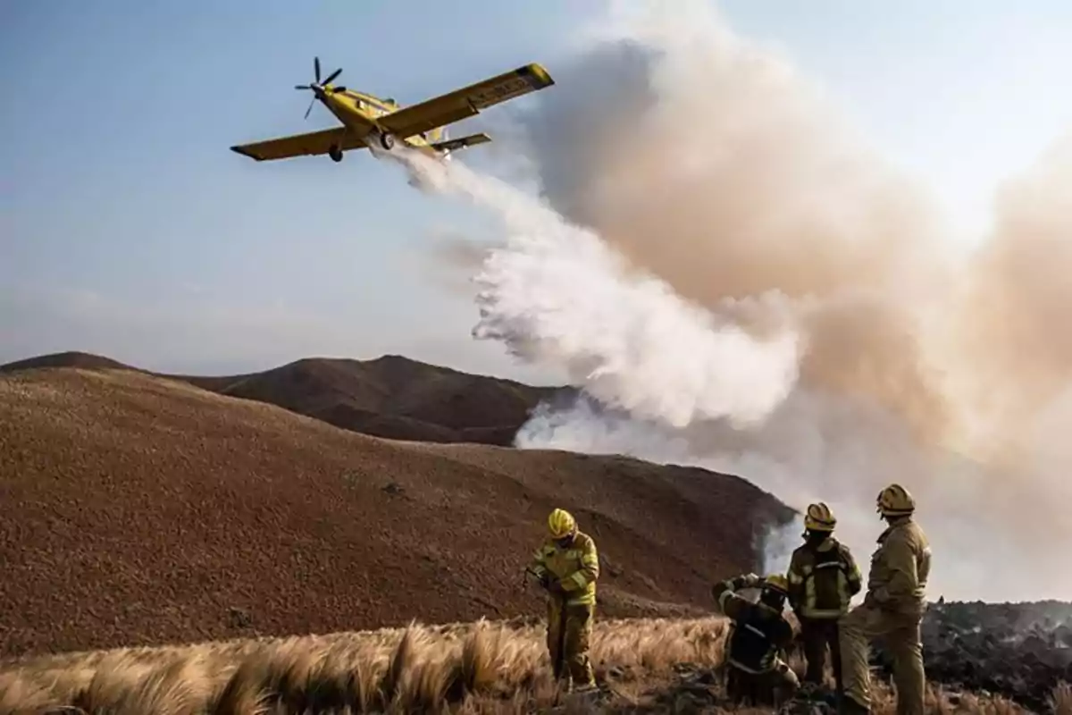 Un avión amarillo arroja agua sobre un incendio forestal mientras varios bomberos observan desde el suelo.