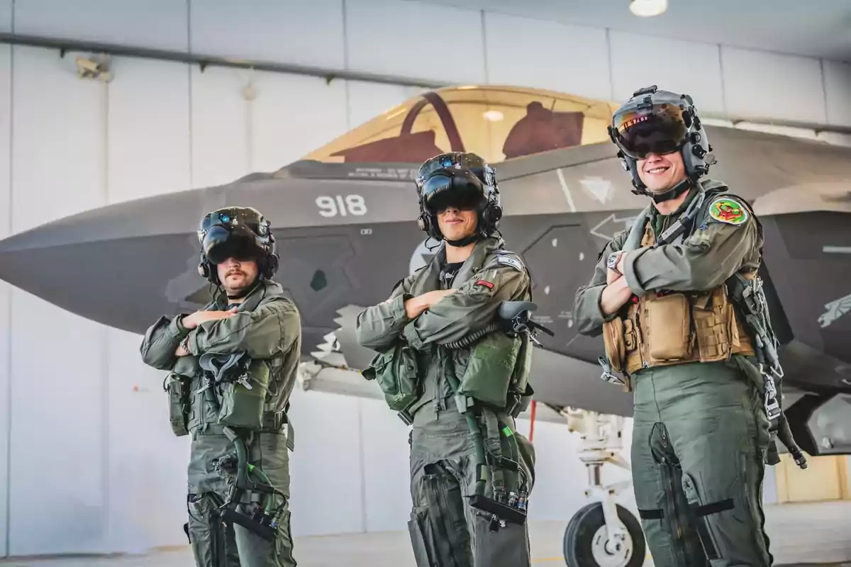 Tres pilotos militares con uniformes y cascos de vuelo posan frente a un avión de combate en un hangar.