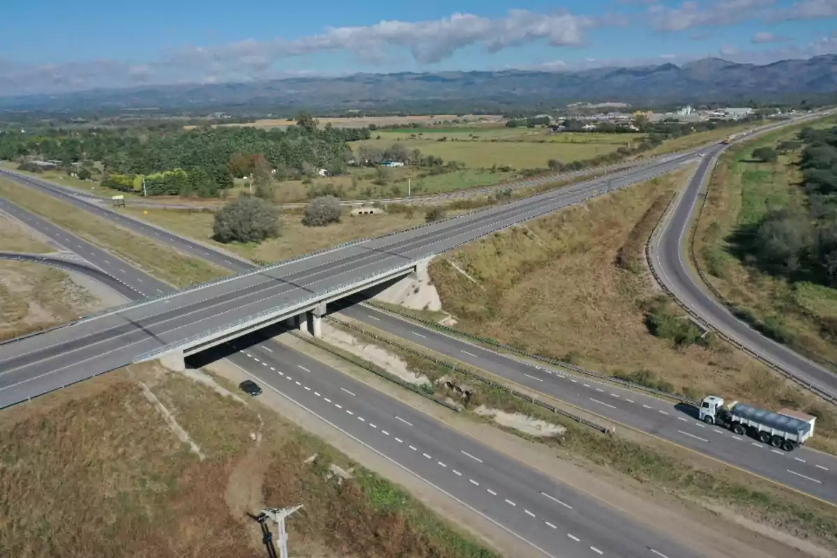 Vista aérea de un cruce de carreteras con un puente y un camión en una de las vías, rodeado de campos y montañas al fondo.