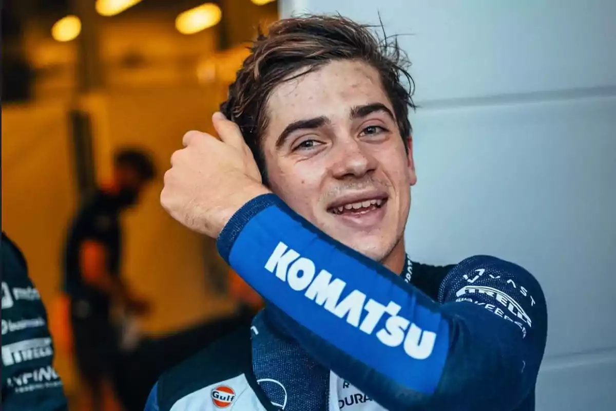 Un joven piloto de carreras sonríe mientras se arregla el cabello, lleva un traje de carreras azul con la marca Komatsu visible en la manga.