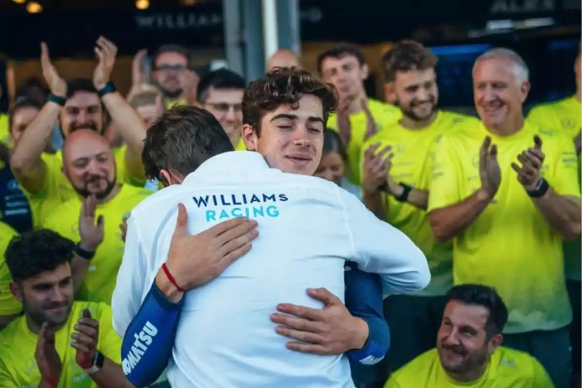 Un grupo de personas aplaude mientras dos hombres se abrazan, uno de ellos lleva una chaqueta con el logo de Williams Racing.