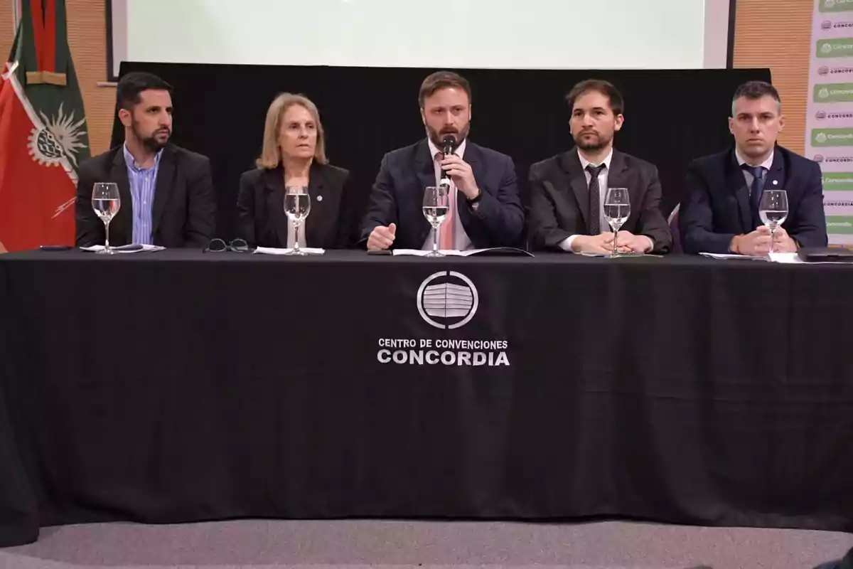 Cinco personas sentadas en una mesa con un mantel negro que tiene el logo del Centro de Convenciones Concordia, con micrófonos y copas de agua frente a ellos.