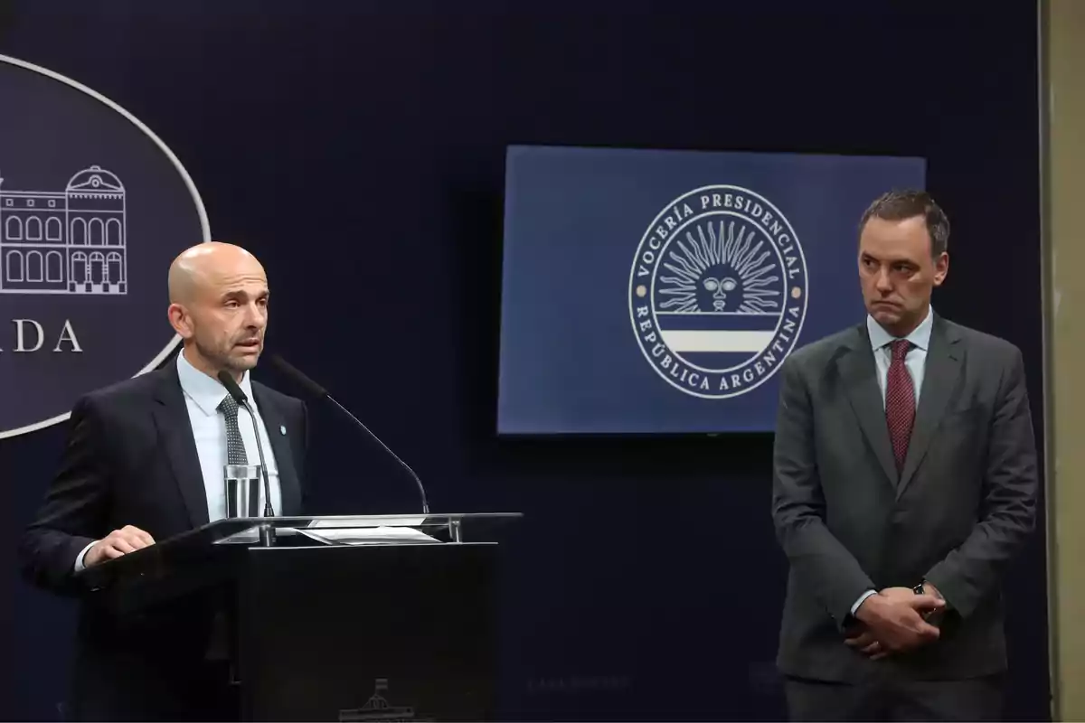 Dos hombres en trajes formales durante una conferencia de prensa en la Casa Rosada con el logo de la Vocería Presidencial de la República Argentina en el fondo.