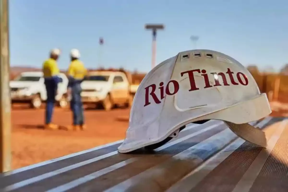 Un casco de seguridad con el logo de Rio Tinto en primer plano y dos trabajadores con cascos al fondo en un sitio de construcción.