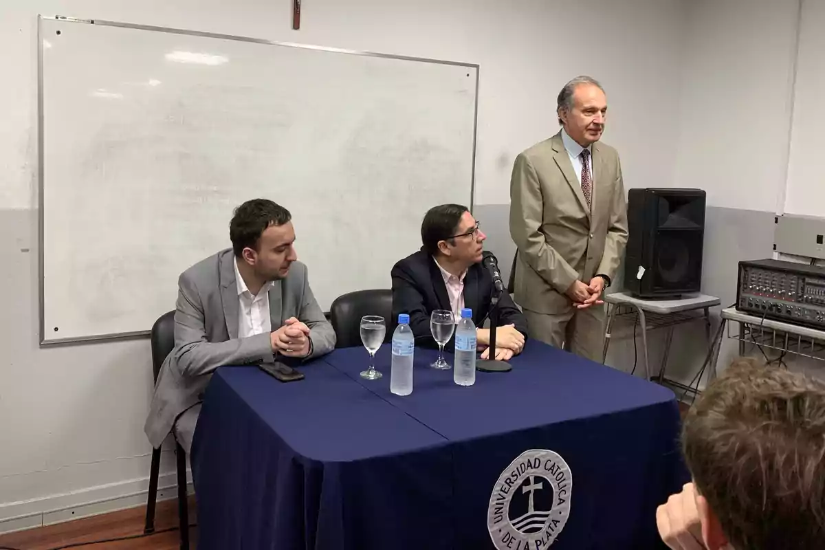 Tres hombres en una sala de conferencias, dos sentados en una mesa con un mantel azul y uno de pie, con una pizarra blanca de fondo y equipo de sonido al lado.