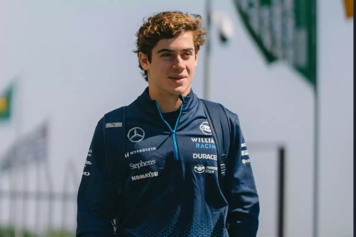 Un hombre joven con chaqueta azul de Williams Racing y mochila sonríe mientras camina al aire libre.