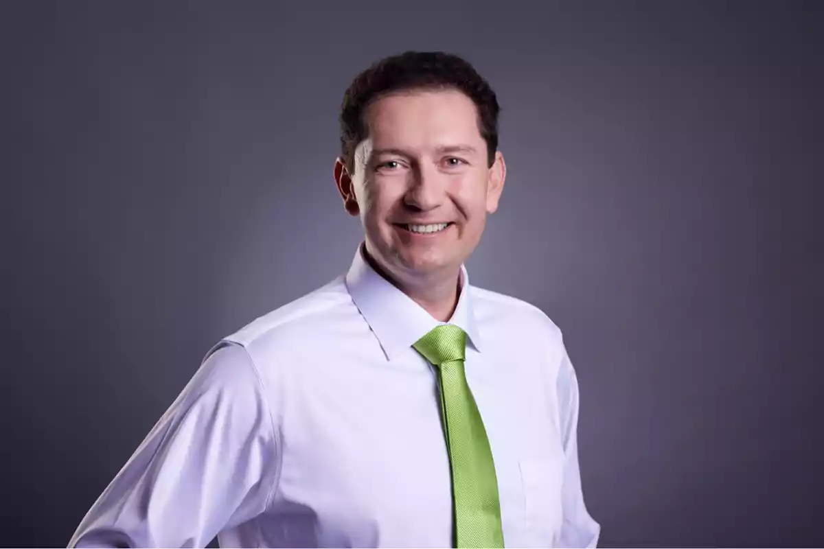 Hombre sonriendo con camisa blanca y corbata verde sobre fondo gris.