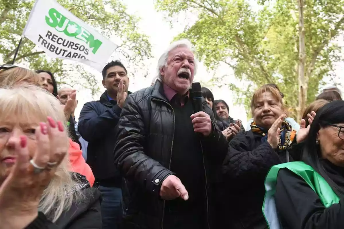 Un hombre mayor con chaqueta negra sostiene un micrófono mientras habla apasionadamente en una manifestación rodeado de personas que aplauden y una bandera que dice "SUOEM Recursos Tributarios" en un entorno al aire libre con árboles.