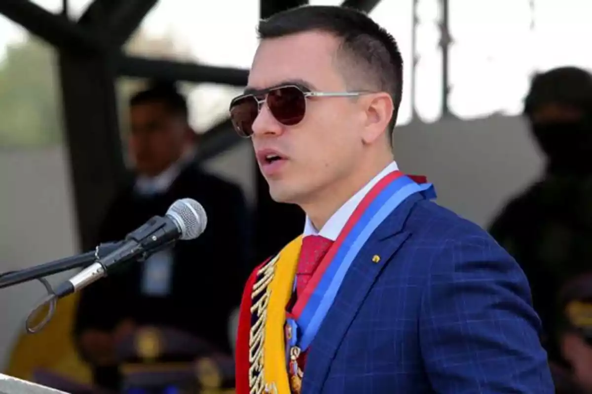 Un hombre con gafas de sol y traje azul habla frente a un micrófono, llevando una banda y medallas.