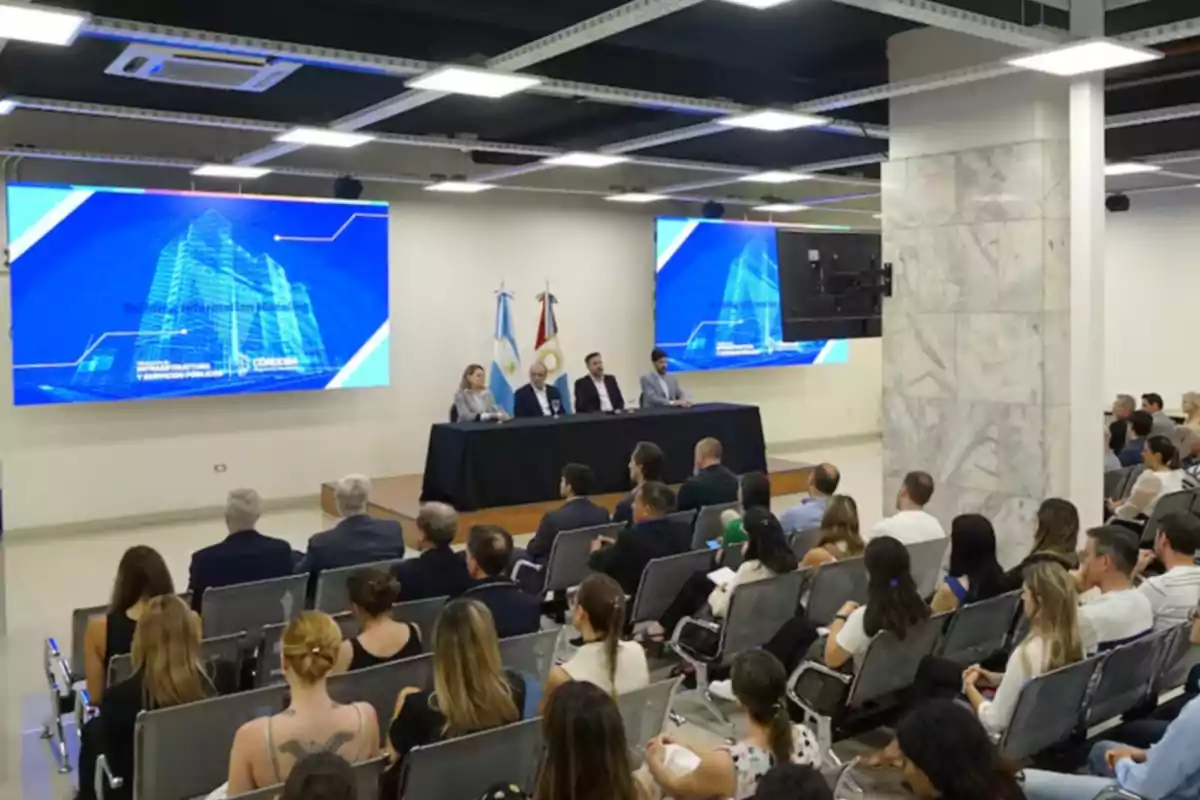 Una conferencia con cuatro personas sentadas en una mesa frente a una audiencia en un salón con pantallas grandes mostrando gráficos de edificios y banderas en el fondo.