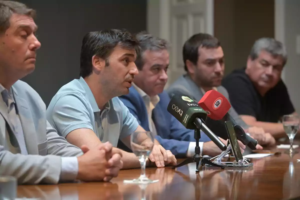 Un grupo de hombres sentados en una mesa durante una conferencia de prensa con micrófonos frente a ellos.